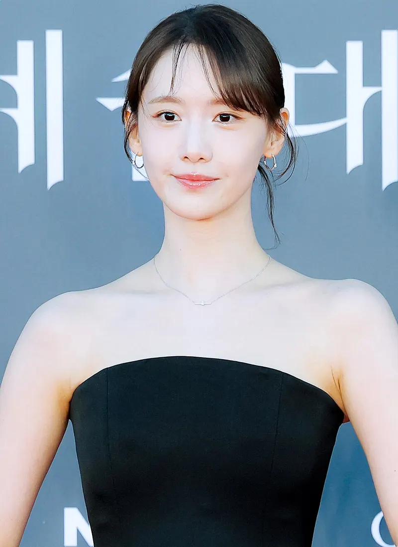 Yoona bergaun hitam tanpa tali berdiri di depan latar bertuliskan huruf Korea