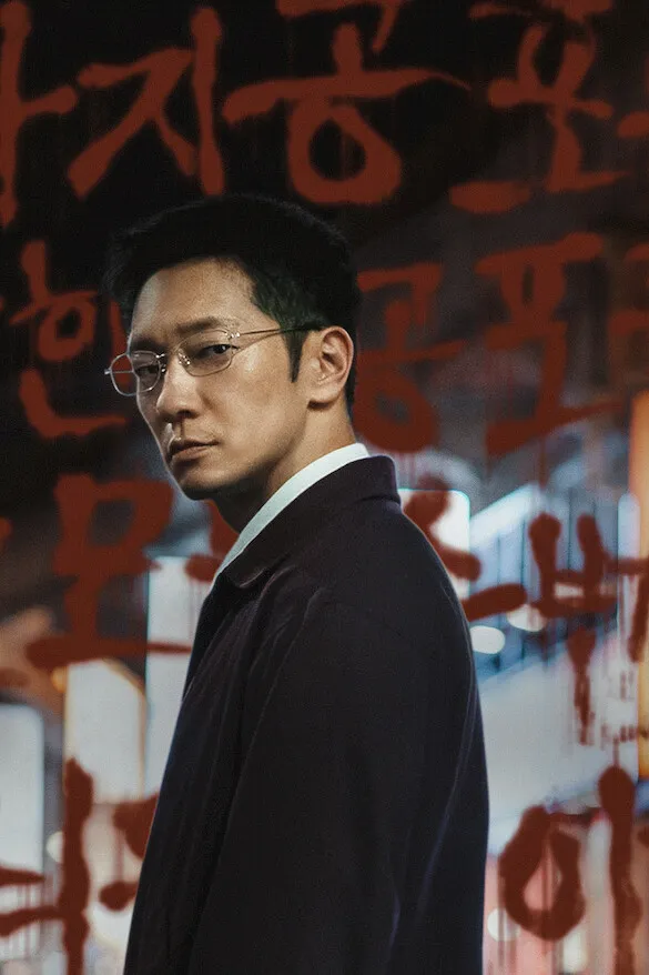 Poster resmi The Blue Road (2026), potongan key art Netflix: Son Suk-ku berkacamata dan bermantel gelap menoleh ke arah kamera, berlatar tulisan Korea merah seperti darah dan lampu kota yang kabur